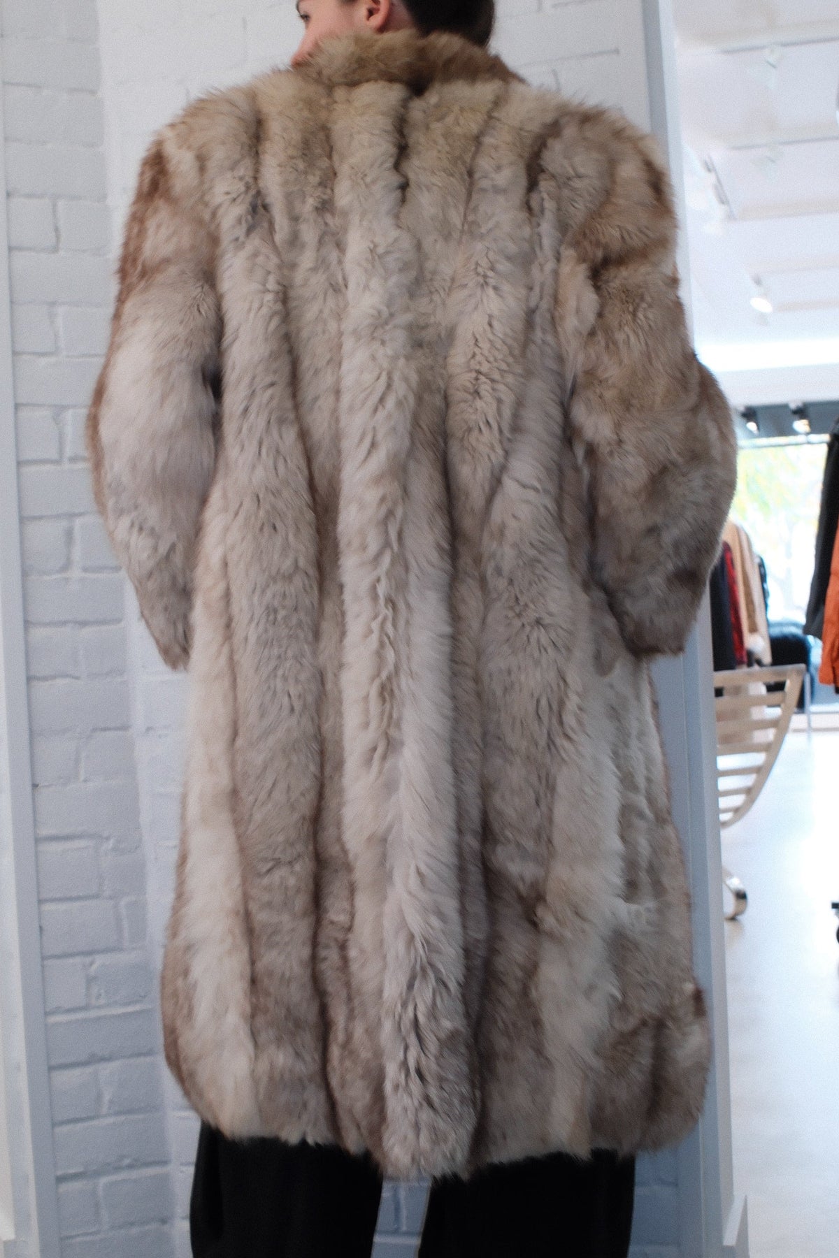 A person wearing a long, plush faux-fur or fur coat in a light taupe-gray color, paired with black pants, standing in an indoor room with a white brick-patterned wall.