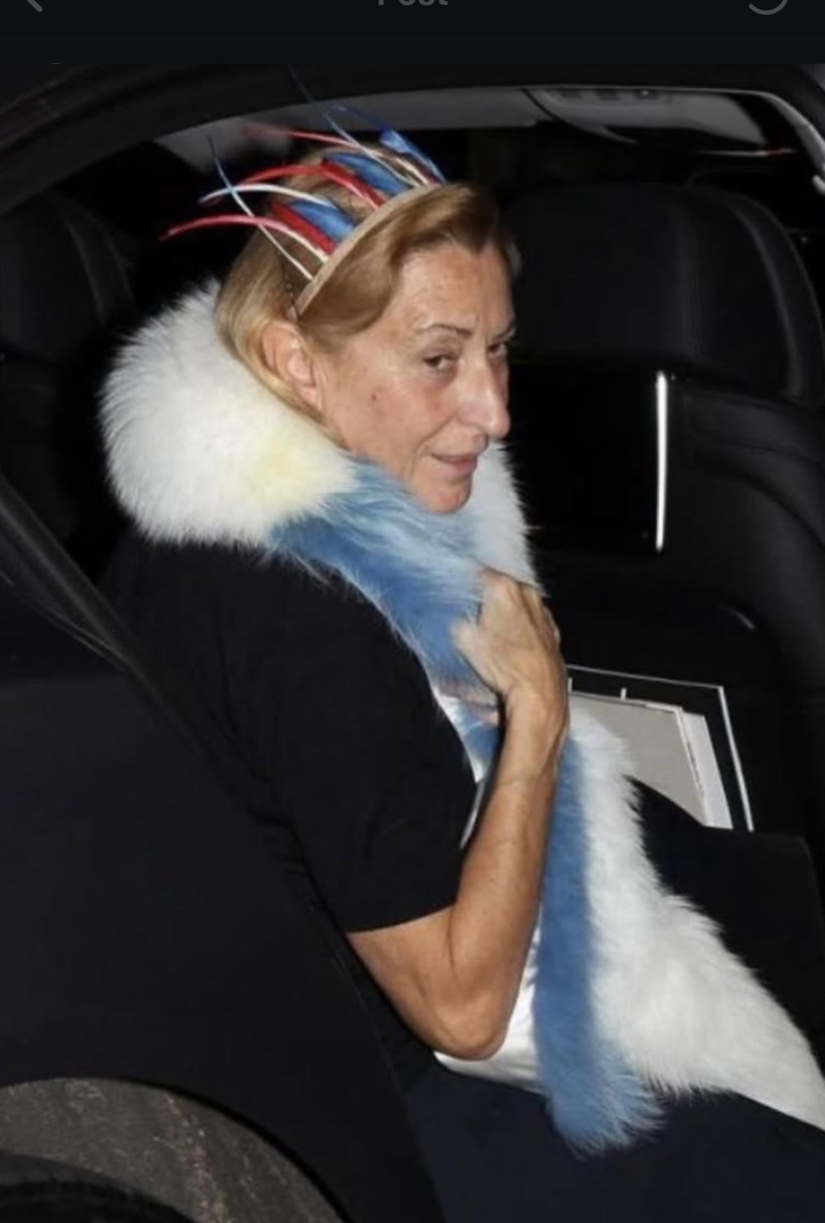 Miuccia Prada wearing a colorful headpiece and fur coat sitting inside a vehicle.