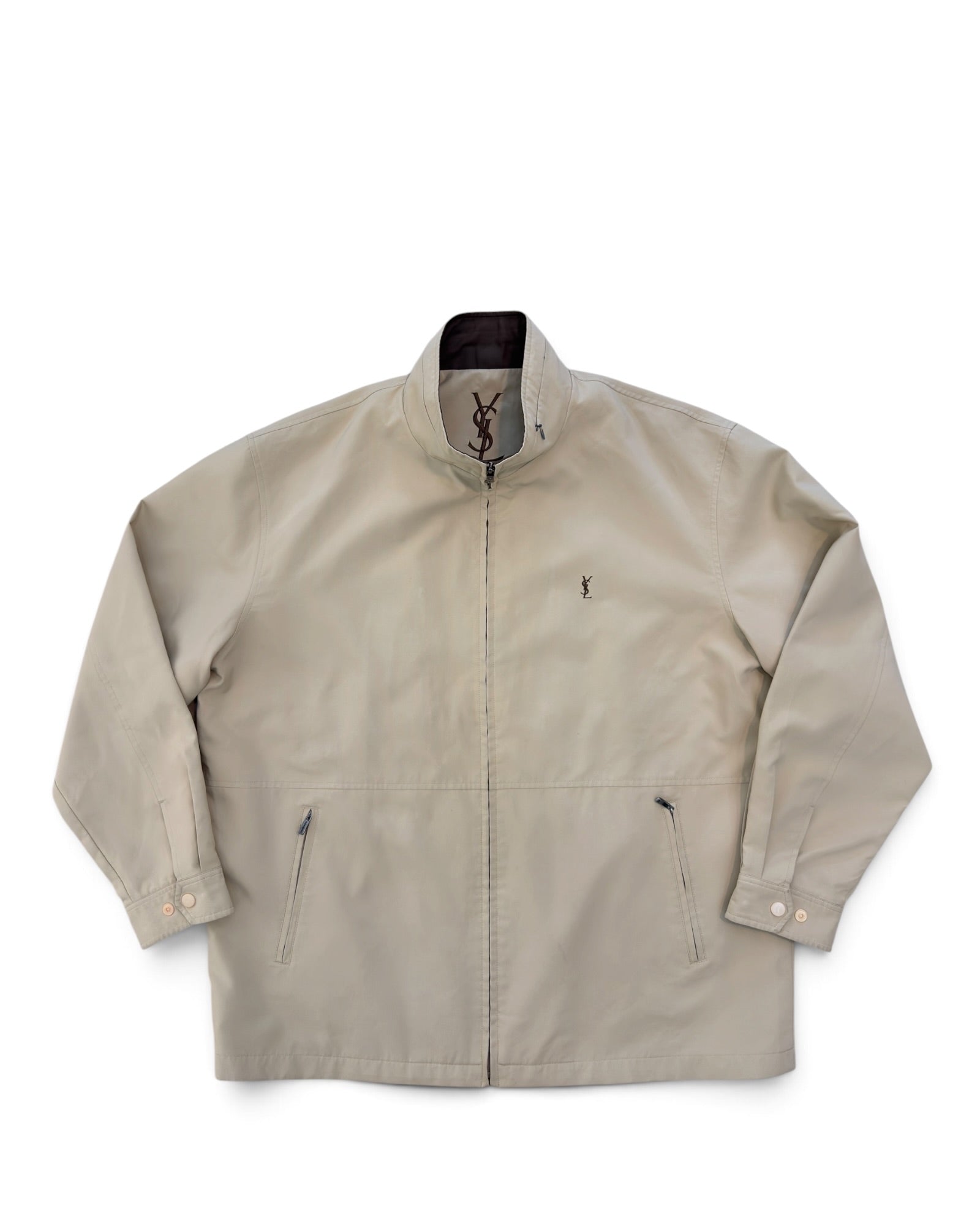 A light beige zip-up jacket with a stand collar, featuring a small embroidered YSL logo on the left chest and a YSL logo printed inside the collar, displayed on a clean white background.