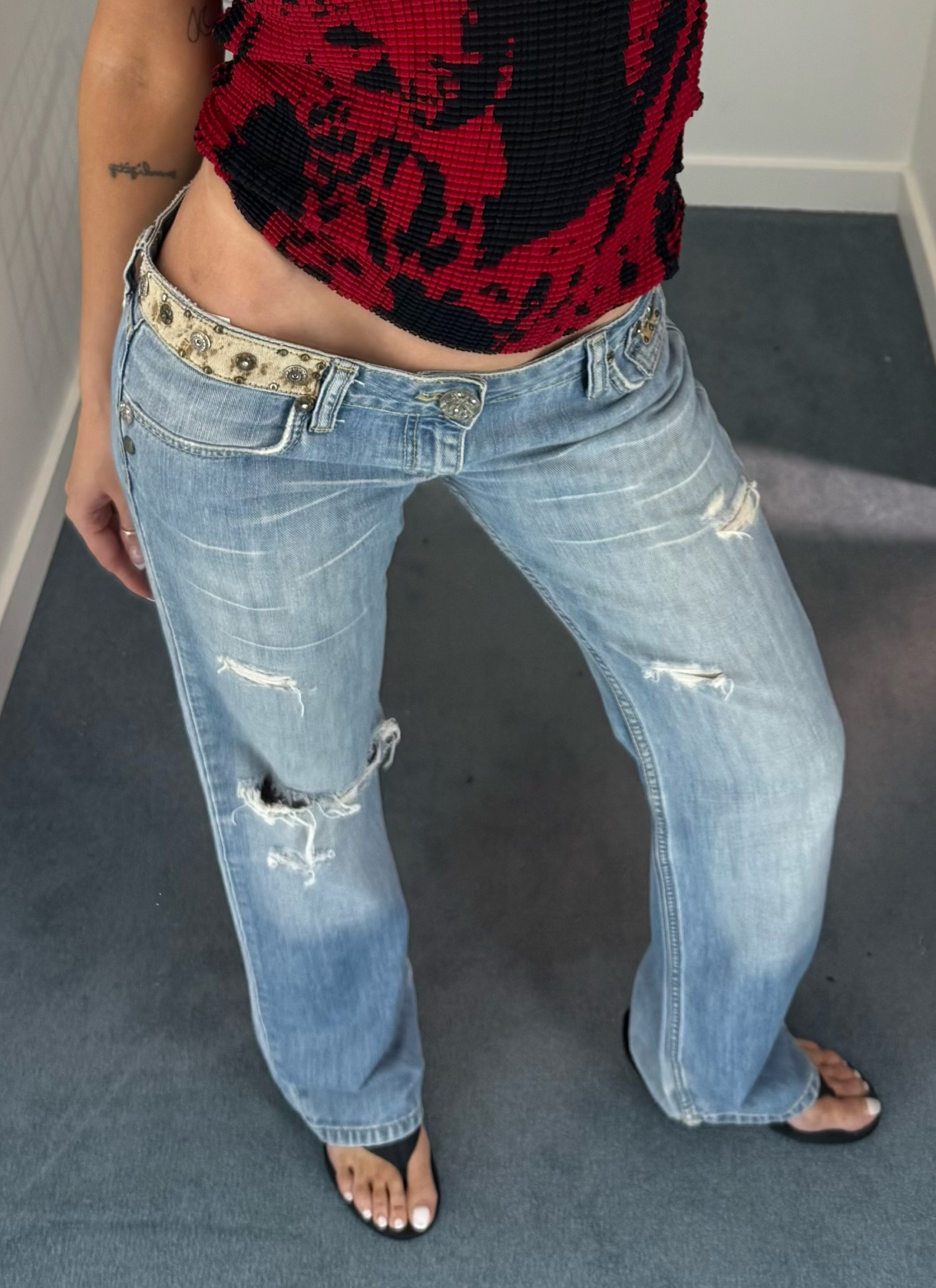 A woman takes a mirror selfie in a fitting room wearing a red and black patterned short-sleeve crop top, distressed light-wash blue jeans, and flat sandals.