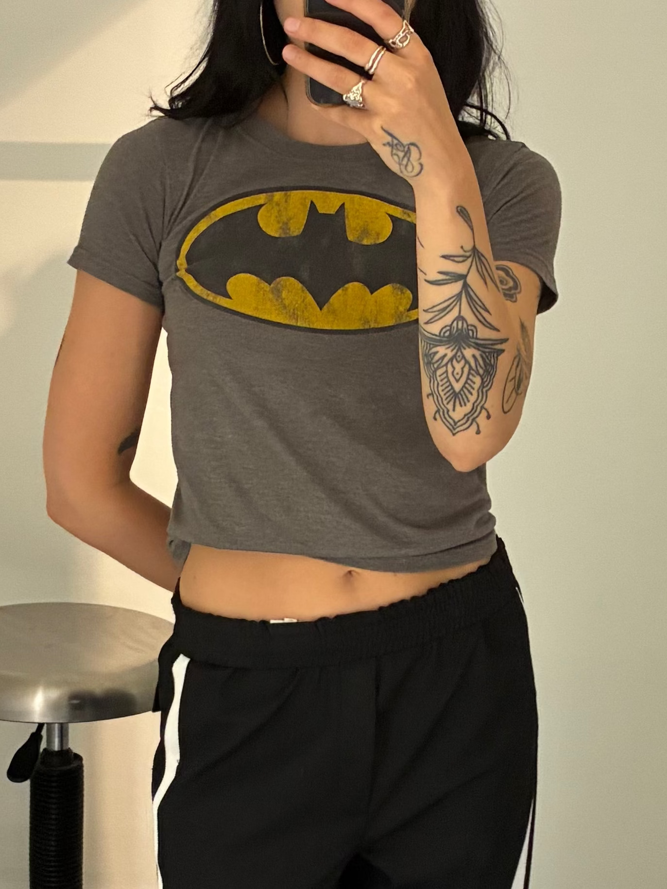 A person taking a mirror selfie in a fitting room wearing a gray cropped t-shirt with a yellow Batman logo and black track pants with white side stripes.