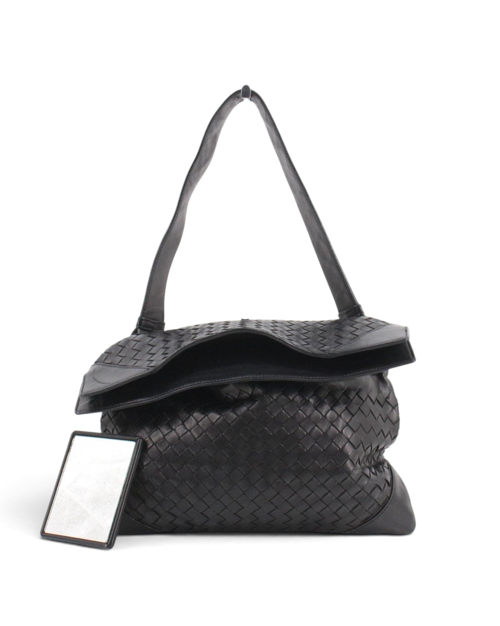 A black woven leather bottega shoulder bag with a flap top and long strap, shown on a plain white background with a small rectangular mirror placed beside it.